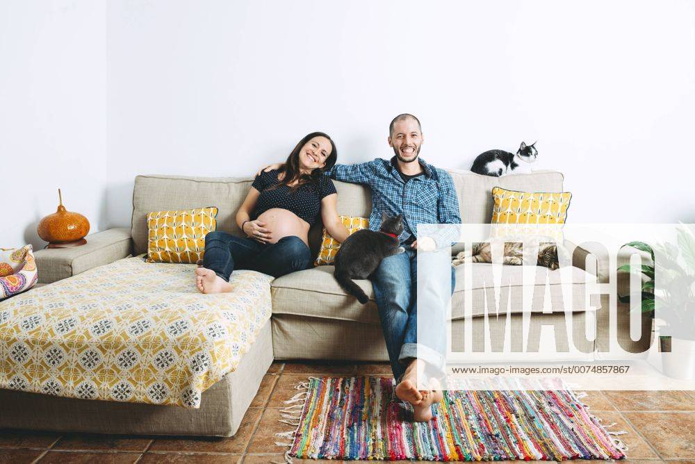 Happy couple expecting a baby sitting on couch at home with three cats model released Symbolfoto property released GEMF01205