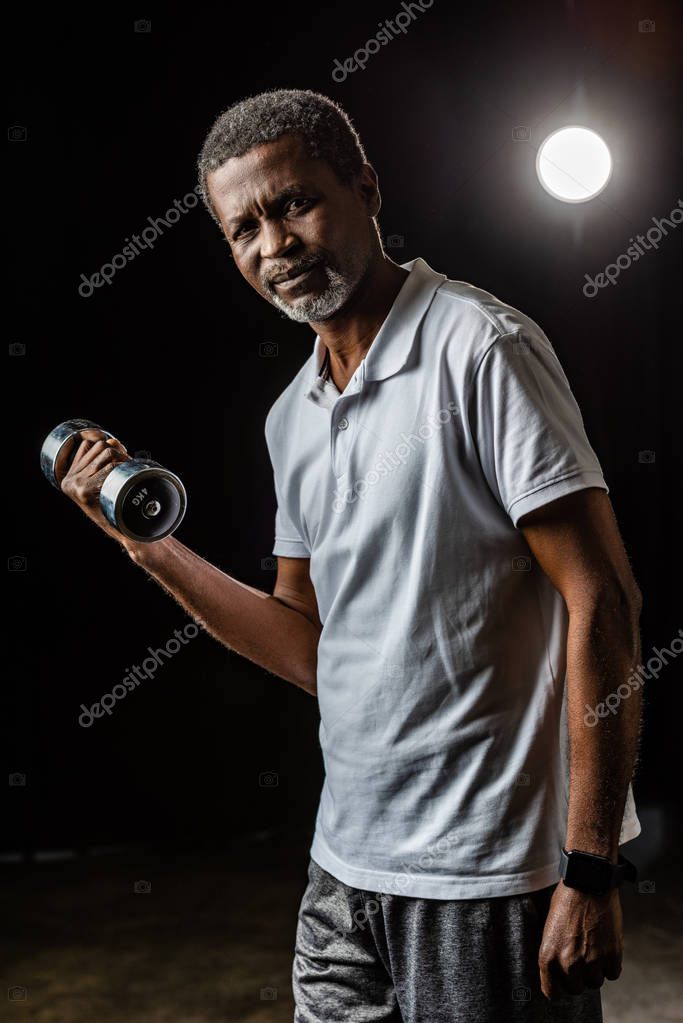 Mature sportsman exercising with dumbbell on black with spotlight