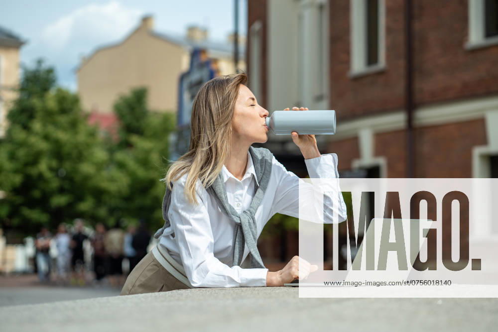 IMAGO Creative, Jahreszeit Sommer, Hitze Happy woman with laptop outside drinking water from bottle, coping with dehydration, thirst. Businesslike girl using computer, working outside, enjoying fresh air, warm weather. Satisfied smiling woman replenishes balance of fluids in body by drinking water with pleasure at hot summer day. ,model released, Symbolfoto