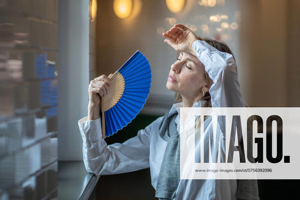 IMAGO Creative, Jahreszeit Sommer, Hitze Woman suffering from heat stuffy air in office without air conditioning fanned herself with hand fan Woman, tired of stuffiness at work, suffering from heat or headache, sweltering stuffy air in an office without air conditioning, stepped out into hallway, fanning herself with paper fan. ,model released, Symbolfoto