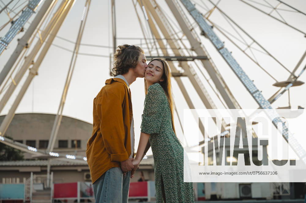 Boyfriend kissing girlfriend standing in front of ferris wheel at amusement park model released, Symbolfoto, ALKF01271