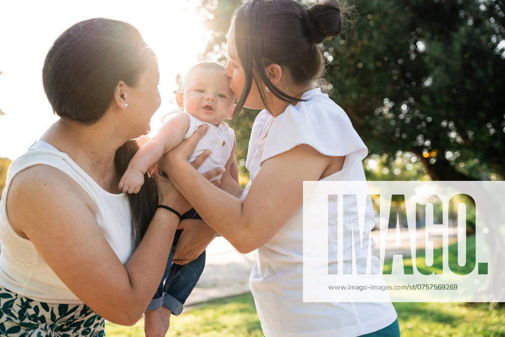A heartwarming scene of a lesbian couple and their infant son outside on a sunny day. The moms are lovingly engaged with their baby, highlighting the warmth and joy of a modern family. Model Released ManuelRuiz_ID23737_688688_008