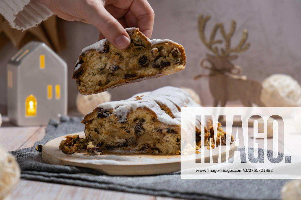 Female hand taking slice of Christmas Stollen in Christmas lights and decoration. Traditional sweet fruit loaf with icing sugar festive pastry dessert. Holiday baking food Female hand taking slice of Christmas Stollen in Christmas lights and decoration. Traditional sweet fruit loaf with icing sugar festive pastry dessert. Holiday baking food concept.