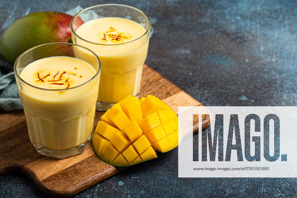 Gesundes indisches Ayurveda-Getränk Mango Lassi in zwei Gläsern auf rustikalem Betontisch mit frischer reifer geschnittener Mango, gelbes Mischgetränk aus Mangofrucht, Joghurt oder Milchquark und Gewürzen copy space Healthy Indian Ayurveda drink mango lassi in two glasses on rustic concrete table with fresh ripe cut mango, yellow blended beverage made of mango fruit, yoghurt or milk curd and spices copy space