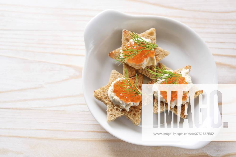 Canapes mit rotem Kaviar auf geröstetem Brot in Sternform mit Sahne und Dillgarnitur auf weißem Teller, heller Holztisch, Kopierraum, Blickwinkel von oben, gewählter Fokus Canapes with red caviar on toasted bread in star shape with cream and dill garnish on a white plate, light wooden table, copy space, high angle view from above, selected focus