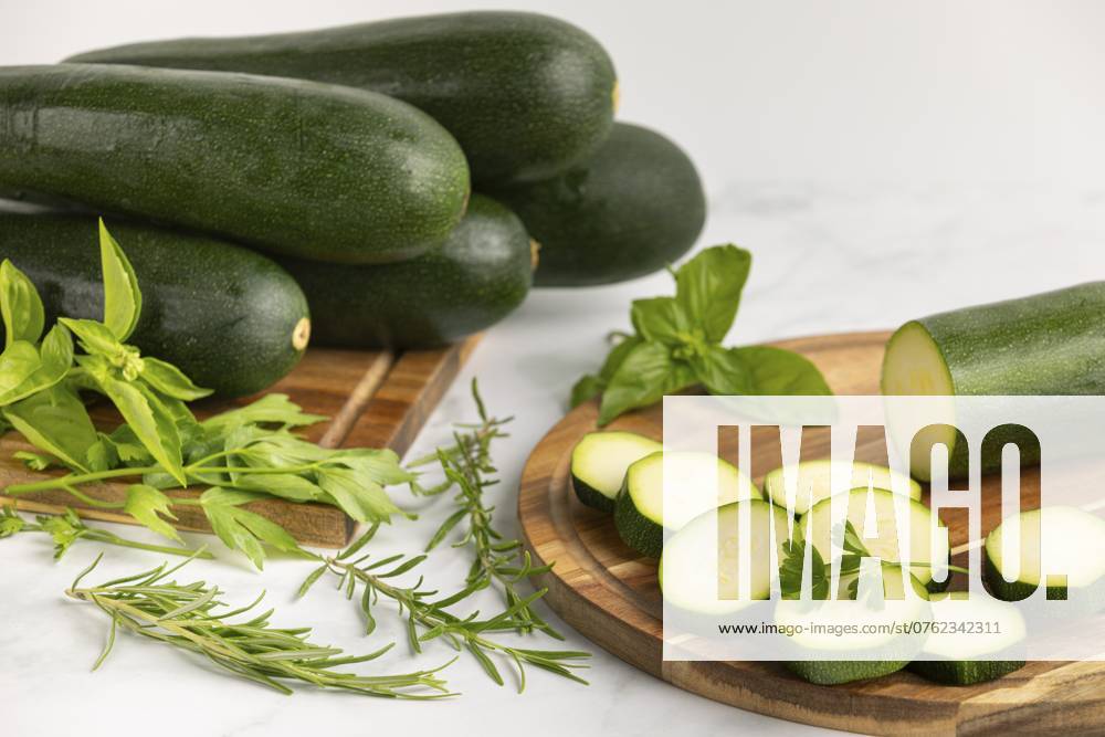 Sliced courgettes on a wooden board, with stacked courgettes and various fresh herbs next to them