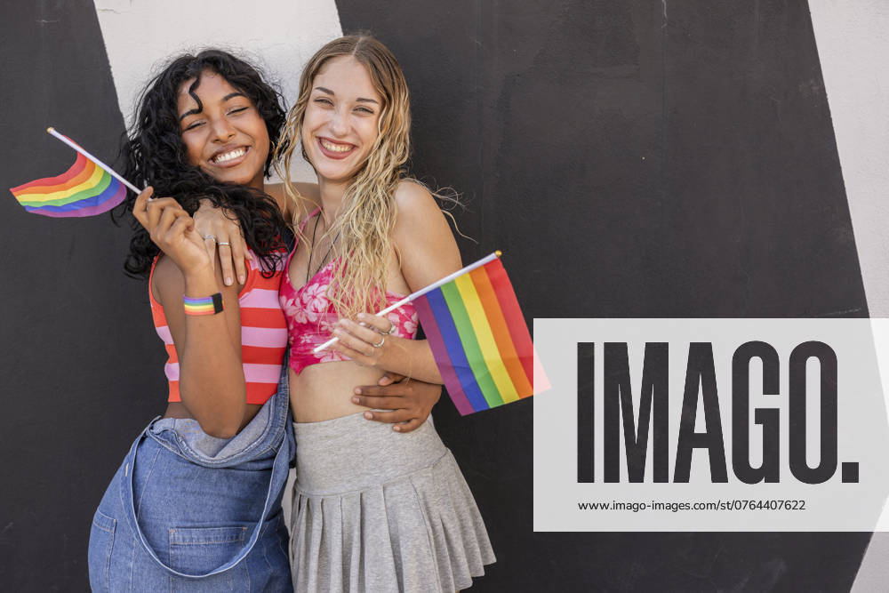 A joyful multiracial lesbian couple embraces while holding LGBTQIA+ pride flags against a contrasting black and white background, symbolizing love and diversity Model Released JoseCarlosIchiro_ID23778_690111_007