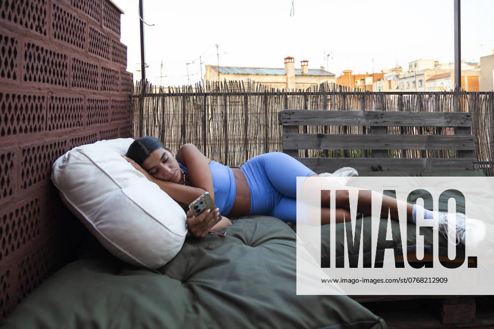 Woman lies comfortably on outdoor bed while operating her smartphone at sunrise in Barcelona, looking at the screen Model Released Almuzambrana_ID23844_692095_008