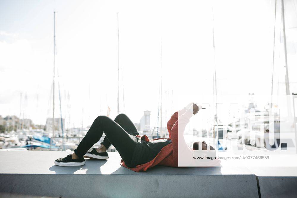 Young man lying on a wall at the marina using cell phone model released Symbolfoto JRFF01171