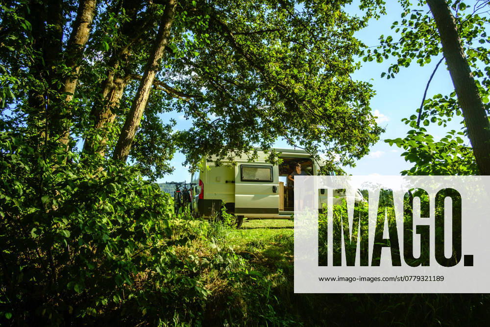 A woman stands in the sliding door of a campervan in an idyllic summer setting in a meadow between trees, Vosges, France Traveling in a campervan