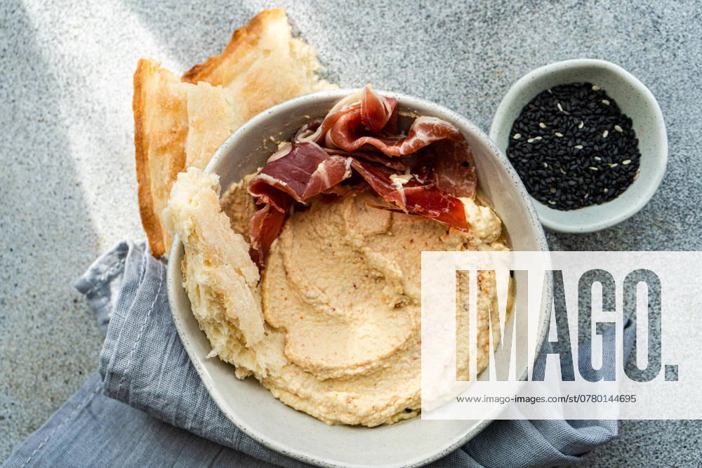 IMAGO Creative, Food, Tapas Top view of a ceramic bowl filled with hummus topped with slices of jamon, accompanied by toasted bread and a side dish of black sesame seeds, presented on a textured grey surface. Model Released AnnaBogush_ID24862_720224_007