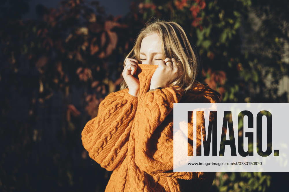 A woman basks in the warmth of a cozy knitted orange sweater, engulfed in sunlight with a backdrop of autumnal foliage, embodying comfort and the essence of seasonal style Model Released HannaYakimenko_ID25074_726416_023