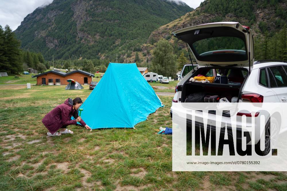An asian woman secures a bright blue tent near her open car trunk in a grassy campsite with scenic mountain views The setup suggests a peaceful outdoor adventure in nature Model Released JuanAlbertoRuiz_ID24637_713881_007