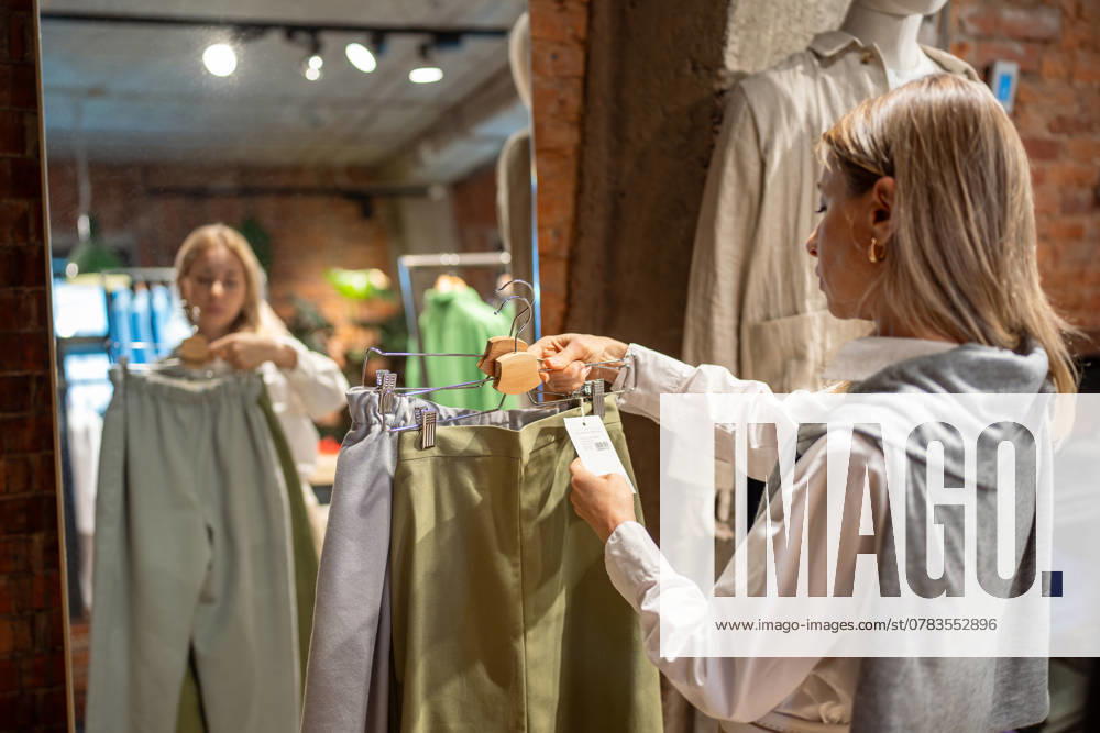 Thoughtful woman shopping for new clothes in store, holds hanger of cotton pants checking price tags Interested middle aged woman shopping for eco outfit, holding hanger of natural organic cotton pants checking price tags in clothing store. Stylish female chooses clothes looks composition of fabric. ,model released, Symbolfoto