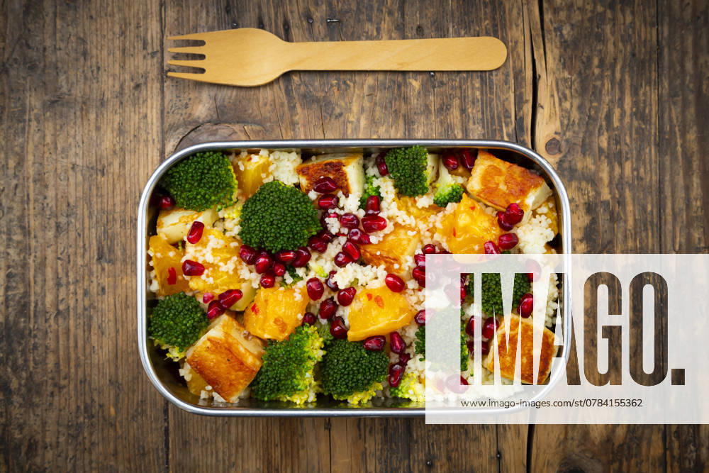 Stainless steel lunch box with couscous, broccoli, halloumi, oranges, chili dressing, and pomegranate seeds, on a wooden table with a wooden fork., LVF09377