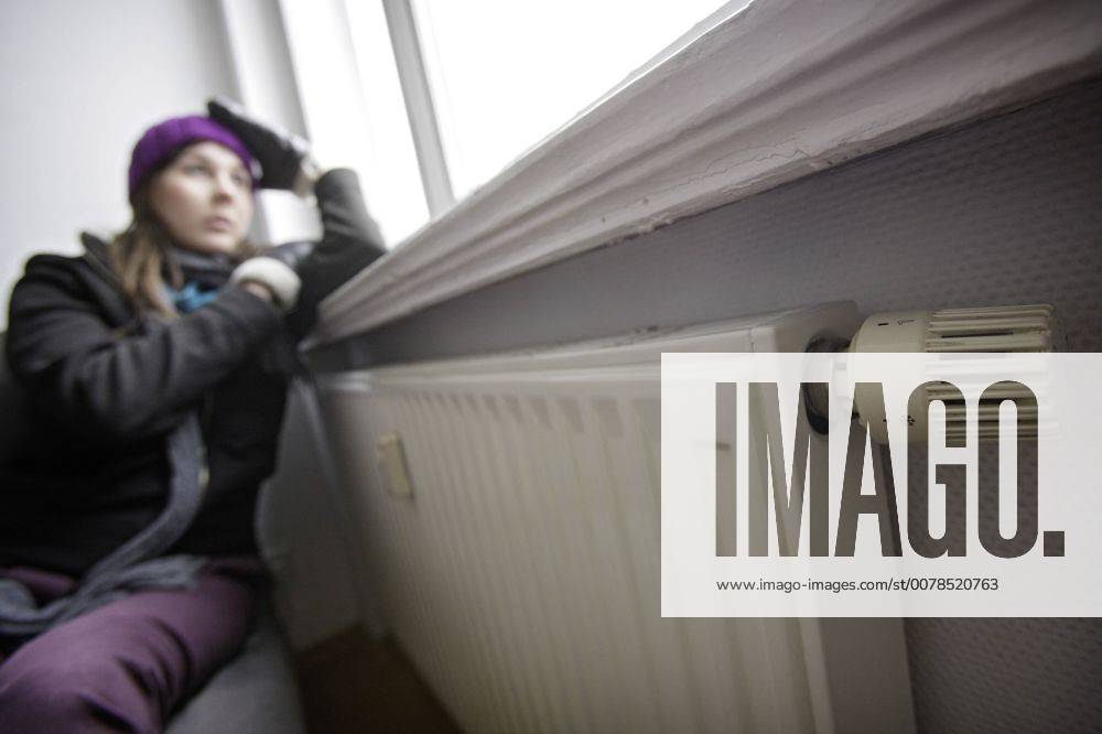 Heizkosten sparen , junge Frau sitzt an der Heizung mit Muetze und Handschuhen . Berlin , Deutschland . Saving heating coasts, young woman at home wearing a cap and gloves . Berlin , Germany . 12.02.2009 , MODEL RELEASE vorhanden , MODEL RELEASED , Berlin DEU Deutschland