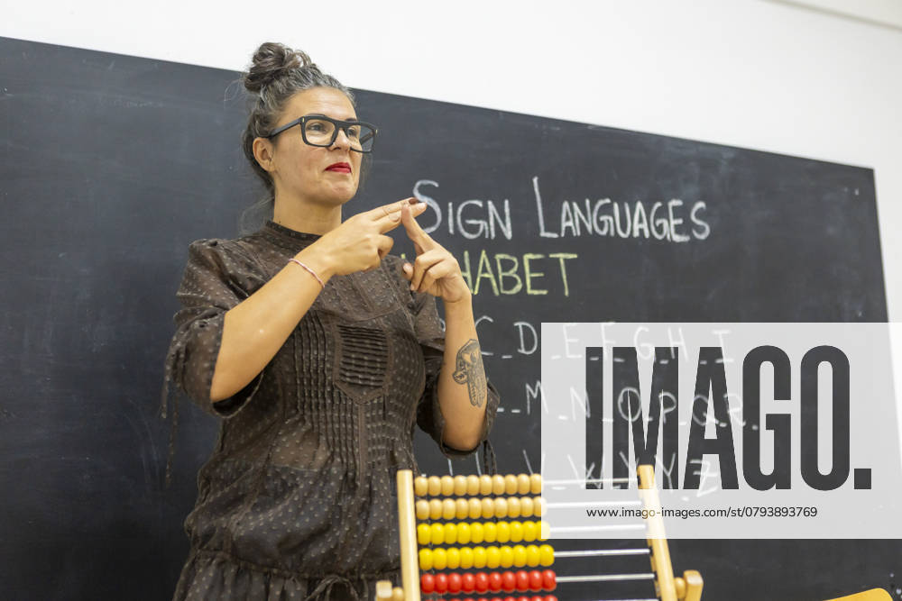 An instructor teaching sign language in a classroom, standing in front of a chalkboard with the alphabet written on it An abacus is visible in the foreground Model Released JuanAizpuru_ID25565_738874_008