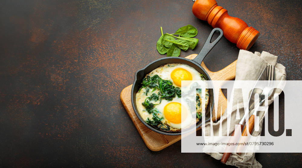 Gesundes Spinat-Omelett in gusseiserner Pfanne zum Frühstück Ansicht von oben Two fried eggs with spinach in a cast iron pan on a wooden board with fresh ingredients, healthy breakfast