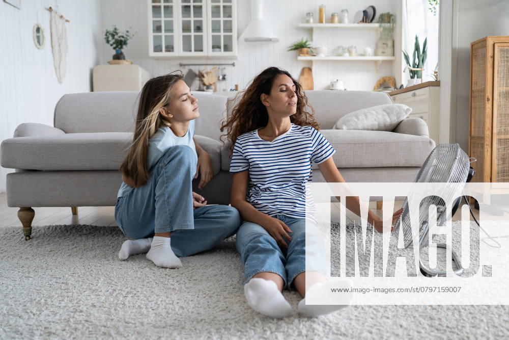 IMAGO Creative, Jahreszeit Sommer, Hitze Liebevolle sorglose Frau Mutter und Teenager-Mädchen sitzt in entspannter Position auf dem Boden im Wohnzimmer in der Nähe von Fan Loving carefree mother and teenage girl sits in relaxed position on floor in living room near fan. Satisfied positive family of woman and daughter spend time at home during abnormally high temperature ,model released, Symbolfoto
