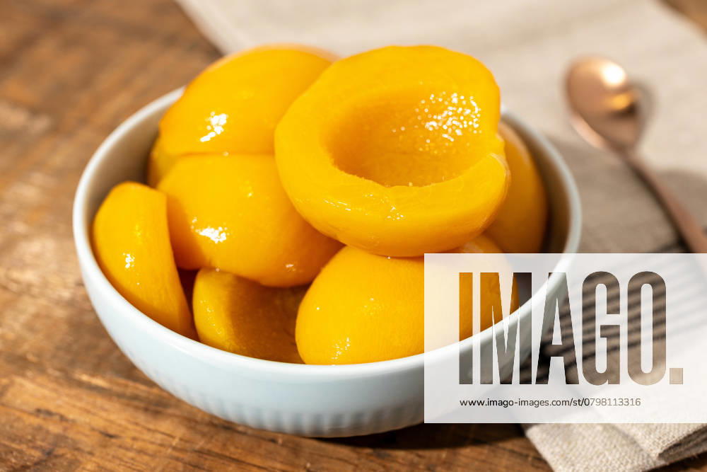 Nahaufnahme einer Schale mit Pfirsichhälften aus der Dose auf einem rustikalen Holztisch mit Serviette und Löffel Close up of a bowl full of canned peach halves on rustic wooden table with napkin and spoon