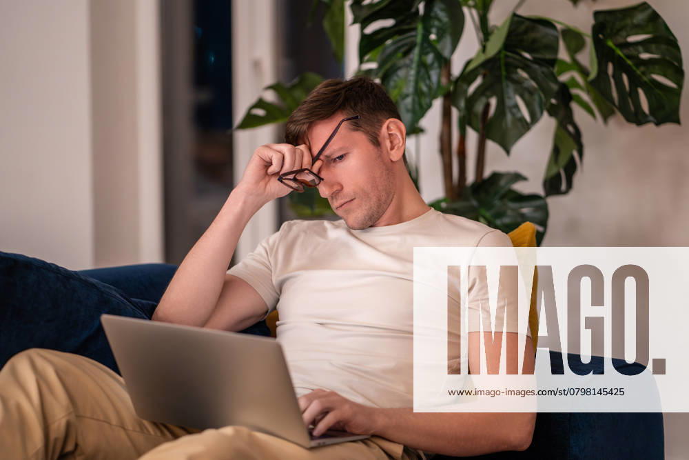 Müder, schläfriger Mann, der sich die Augen reibt und am Abend auf der Couch sitzt. Tired sleepy man freelancer workaholic rubbing eyes working on laptop sitting on couch in evening at home. Overwork, over stain, hard work, career ambitions concept. Distant, remote job, freelance. ,model released, Symbolfoto