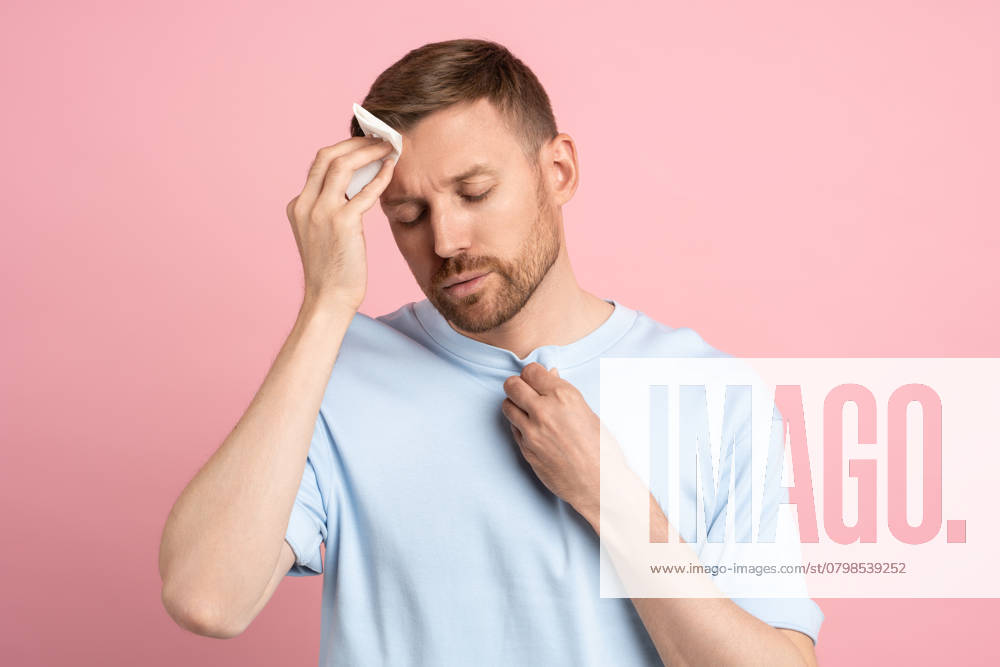 Ein Mann, der unter Hitzestau leidet, wischt sich mit einer Papierserviette den Schweiß von der Stirn, isoliert auf einer rosa Wand Man suffering from heat stuffiness wiping sweat on forehead with paper napkin isolated on pink background. Exhausted overheated bearded male with closed eyes. Summer hot weather, stuffy room concept. ,model released, Symbolfoto