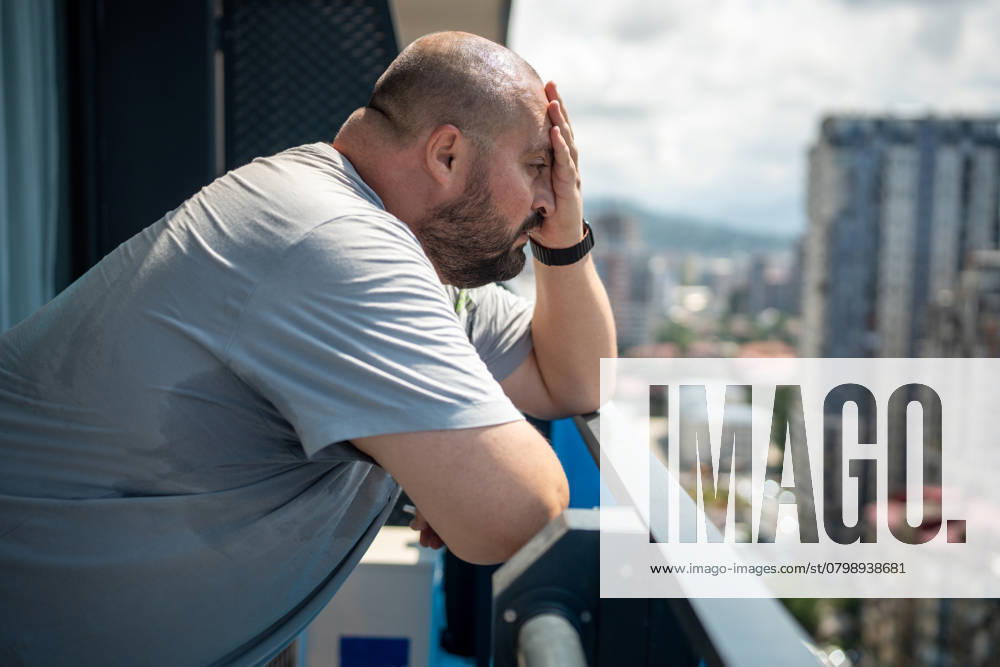 Übergewichtiger Mann, der sich bei sommerlicher Hitze schlecht fühlt, steht auf einem Balkon und schaut auf die Straße. Overweight man feeling bad from heat in summer standing on balcony looking at street in summer. High sweating, hyperhidrosis, obesity, heaviness from hot weather high humidity, excess body weight. ,model released, Symbolfoto