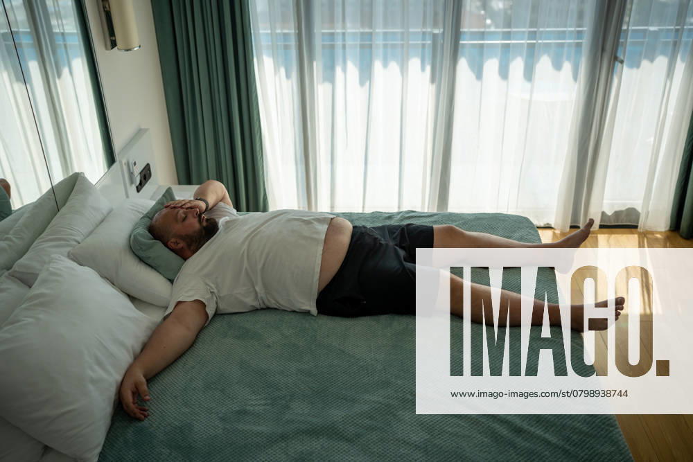 Fauler, erschöpfter, übergewichtiger Mann, der im Bett schläft und sich im Urlaub im Hotelzimmer schlecht fühlt. Lazy overweight man sleeping in bed on vacation in hotel room. Tired exhausted guy with excess weight feel bad in hot summer weather resting in daytime. Heat, high blood pressure, fatigue concept. ,model released, Symbolfoto