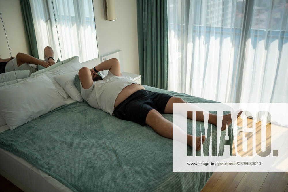 Ein fetter, übergewichtiger Mann stirbt vor Hitze. Faul, fettleibig übergewichtigen Mann liegt auf dem Bett, fühlt sich müde im Zimmer Fat man suffers dying from heat. Lazy, obese overweight young guy lies on bed, suffering from summer abnormal warmth, feels tired, unpleasant discomfort sensations in body, high blood pressure. ,model released, Symbolfoto