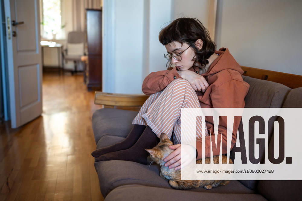IMAGO Creative, Gesellschaft, Leisure Sickness Deprimiert erschöpft Teenager-Mädchen streicheln Katze sitzt auf der Couch zu Hause. Psychisches Problem, Haustier-Therapie. Depressed exhausted teen girl in glasses pyjamas petting cat sitting on couch at home in closed pose. Teenage age crisis, emotional burnout depression, psychological or life problems troubles concept. ,model released, Symbolfoto
