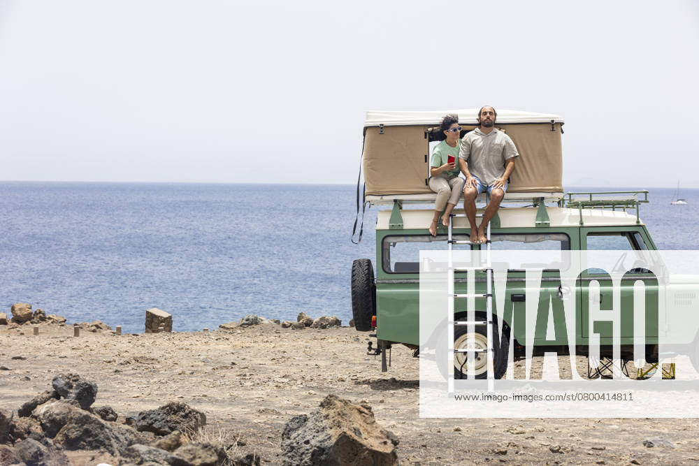 A blind man and a companion sit on the rooftop of a motorhome parked by the sea. They are enjoying the scenic view and feeling the breeze on a sunny day. Model Released JuanAizpuru_ID26099_752916_016
