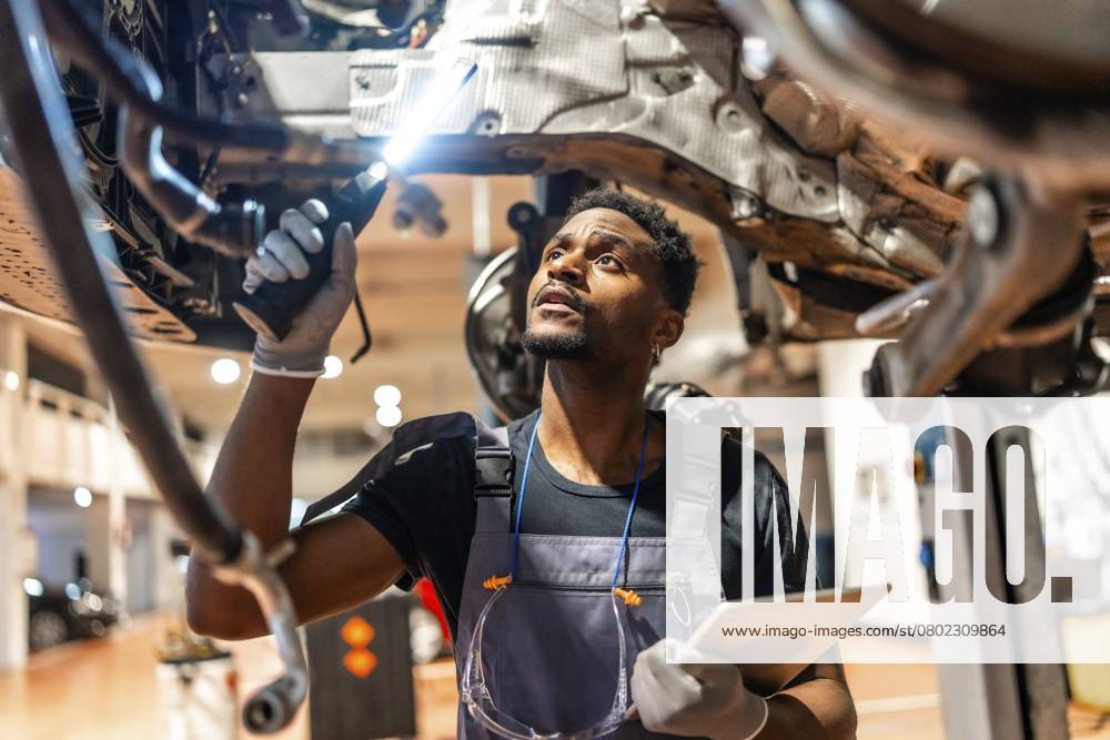 Junger Mechaniker inspiziert den Unterboden eines Autos mit einer Taschenlampe und einem Tablet in einer modernen Autowerkstatt Young mechanic inspecting car undercarriage using flashlight and holding tablet in a modern auto repair shop