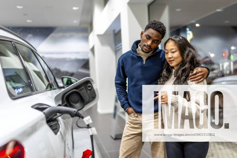Ein multiethnisches Paar untersucht den Ladeanschluss eines Elektrofahrzeugs in einem Autohaus und setzt sich für nachhaltige Mobilität ein Multiethnic couple examining charging port of electric vehicle at car dealership, embracing sustainable transportation