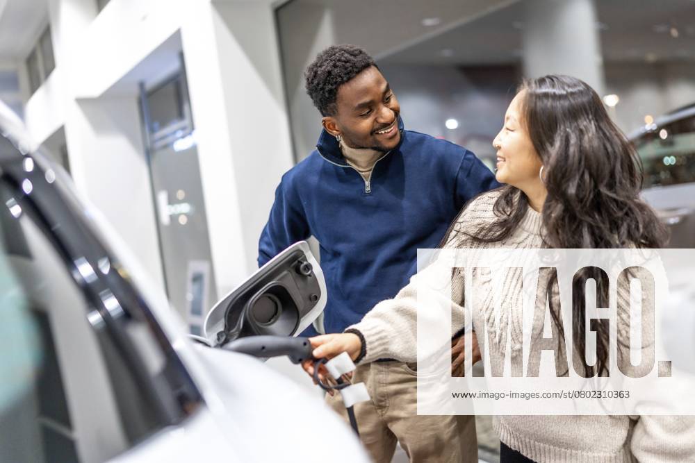 Lächelndes Paar, das die Ladestation eines Elektrofahrzeugs untersucht, während es sich in einem Autohaus über die Möglichkeiten eines nachhaltigen Lebens informiert Smiling couple examining the charging socket of an electric vehicle while exploring options at a car dealership, embracing sustainable living