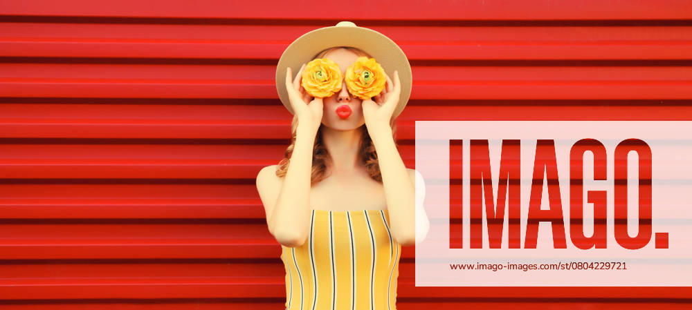 Sommer Porträt der stilvollen jungen Frau bläst Kuss mit gelben Blütenknospen auf rotem Hintergrund Summer portrait of stylish young woman blowing kiss with yellow flower buds looking for something in round straw hat on red background ,model released, Symbolfoto