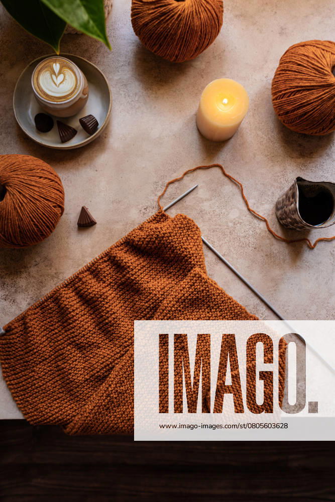 A cozy knitting setup featuring warm brown yarn balls, a knitting project, and a steaming cup of coffee with chocolates, illuminated by a soft candlelight glow. Model Released CintiaViudez_ID26646_768866_002