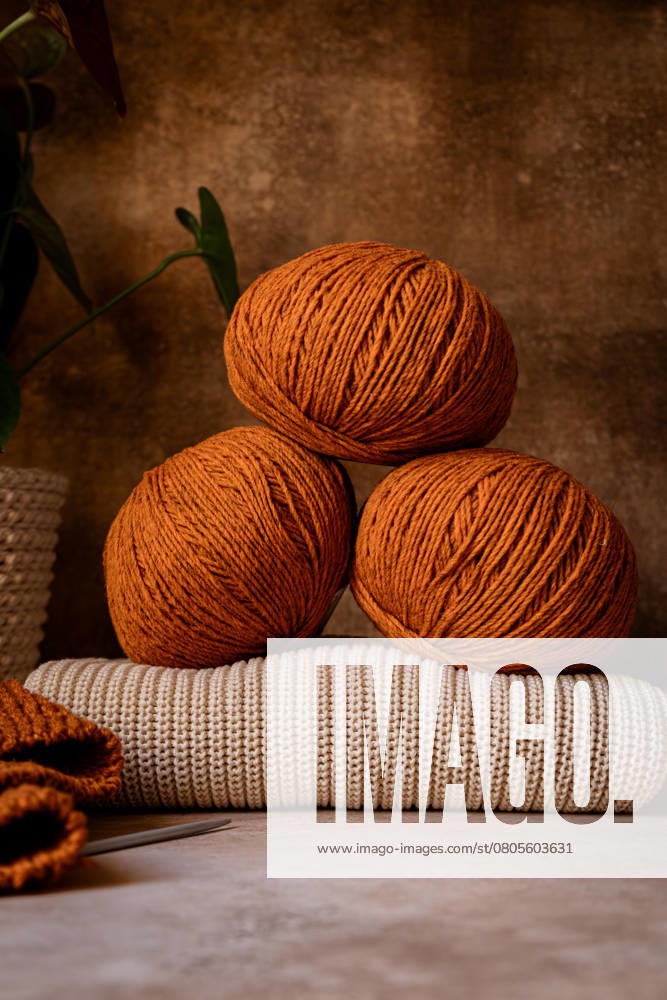Three warm-toned yarn balls sit elegantly on a knitted surface, evoking cozy autumn vibes. The background features natural elements, enhancing the rustic charm. Model Released CintiaViudez_ID26646_768872_005