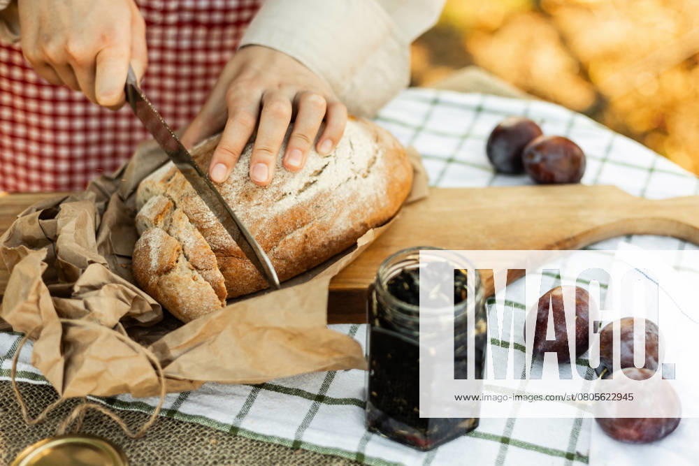 IMAGO Creative, Jahreszeit Sommer, Food, Pflaume Hands slicing rustic bread on a wooden board, surrounded by fresh plums and jam. An inviting scene for preparing delicious homemade canapes in a cozy kitchen setting. Model Released Ptashkimenko_ID26208_756096_006