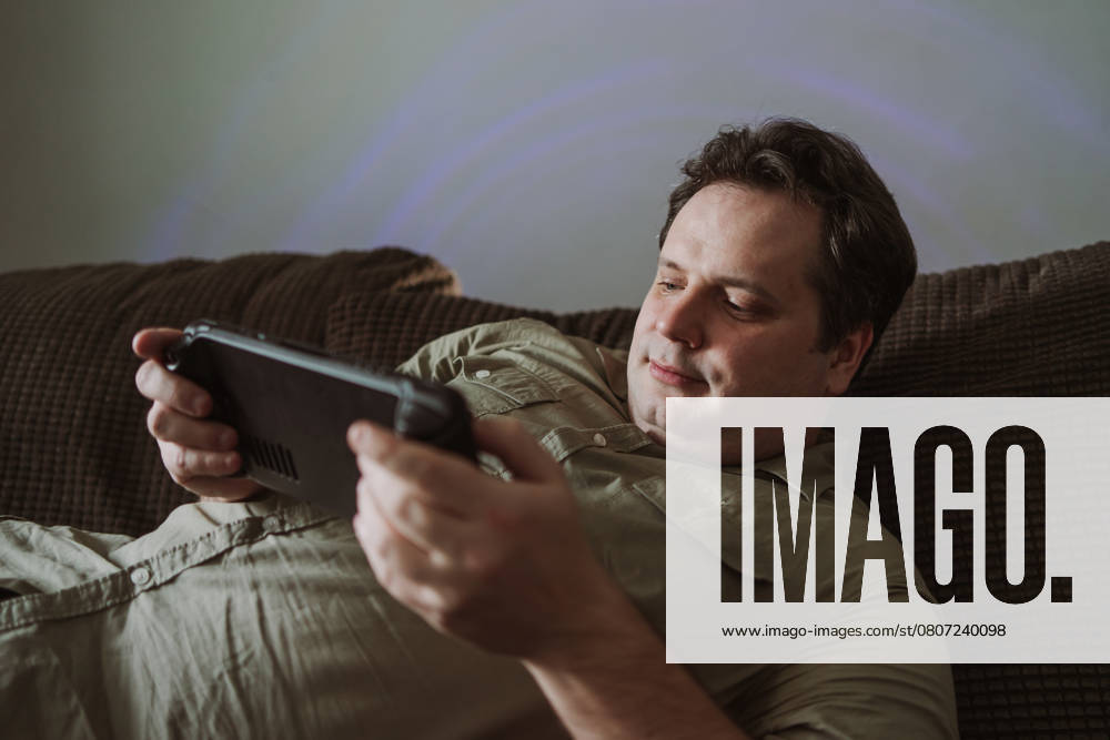 IMAGO Creative, Lifestyle, Gaming A man relaxes on a comfortable couch, focused on playing a game on a hand held device. Casual attire and soft lighting create a cozy home atmosphere. Model Released JenyKulikova_ID26998_779230_022