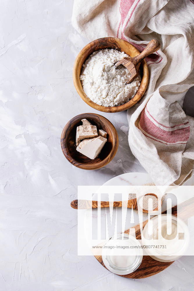 Sourdough for baking bread Rye and wheat sourdough in glass jars, fresh and instant yeast, olive wood bowl of flour for baking homemade bread. With spoon, serving board, kitchen towel over gray concrete background. Top view