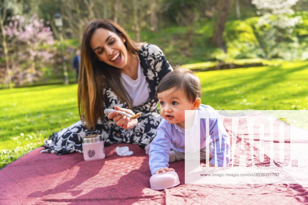 Glückliche Mutter, die ihr Baby mit einem Löffel mit fester Nahrung füttert, sitzend auf einer Decke bei einem Picknick im Park Happy mother feeding her baby solid food with a spoon, sitting on a blanket during a picnic in a park