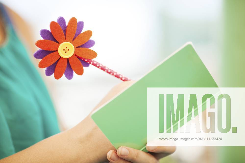 Nahaufnahme einer nicht erkennbaren Frau, die mit einem Bleistift und einer bunten Blume in einem Notizbuch mit einfarbig grünem, pastellfarbenem Umschlag schreibt Close Up of Unrecognizable Woman Writing with Pencil Topped with Colorful Flower in Notebook with Plain Green Pastel Colored Cover