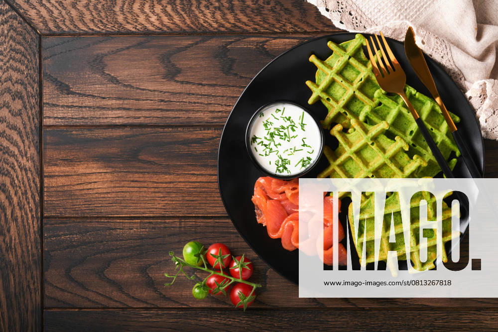 Grüne belgische Waffeln. Spinat-, Bärlauch- oder Pesto-Waffeln mit rotem Lachs und Sahnesauce auf schwarzem Teller vor dunklem, rustikalem Holzhintergrund. Leckeres Frühstück, Snack, Brunch. Ansicht von oben. Attrappe Green Belgian waffles. Spinach or wild garlic or pesto waffles with red salmon and cream sauce on black plate over dark rustic wooden background. Delicious breakfast, snack, brunch. Top view. Mock up (License=RF)