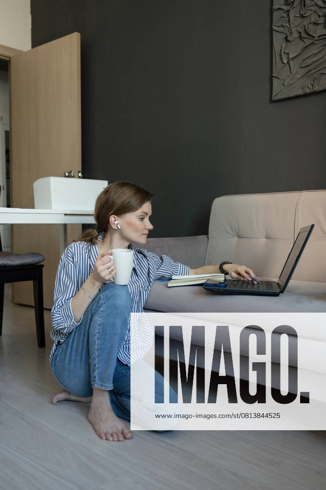MR_ID27929_20617_TatyanaRangulova.jpg, PR_ID27929_10891_Cat.jpg, PR_ID27929_10892_Room.jpg A woman sitting on the floor by a sofa, working on a laptop. She holds a coffee mug and listens with earbuds. The atmosphere is cozy and relaxed at home., Property released Model Released LiliyaSolopova_ID27929_802099_032