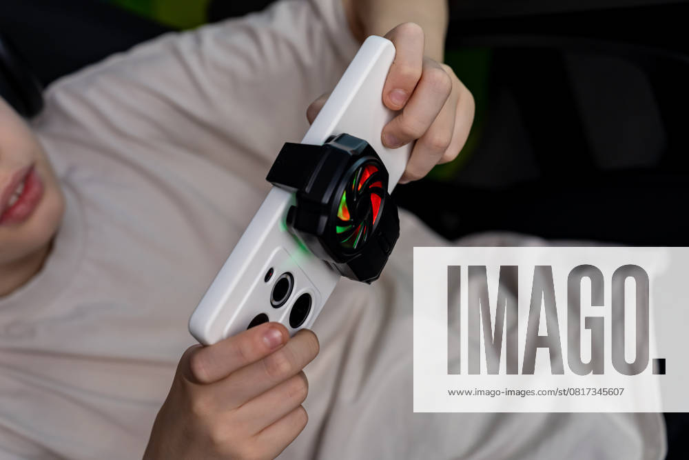 IMAGO Creative, Lifestyle, Gaming Teen boy intensely focused on mobile gaming, holding a smartphone with a cooling fan. The setup enhances gameplay experience and performance during intense sessions Model Released Ikvyatkovskaya_ID28180_830165_031