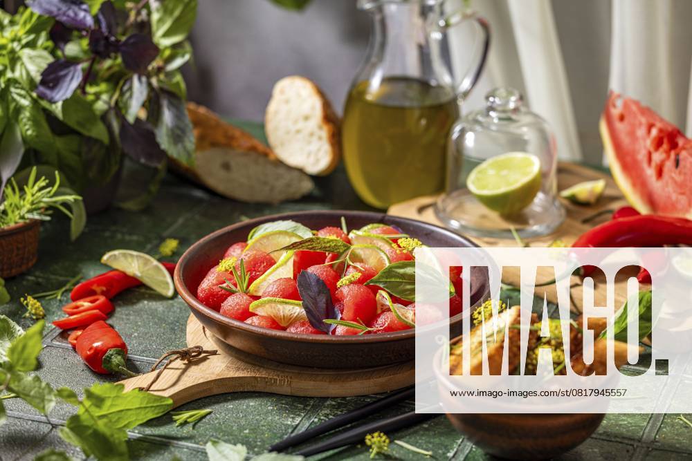 Pikanter Salat aus frischen Wassermelonenkugeln mit Basilikum, Limette, Rosmarin und Fenchel, mit Olivenöl beträufelt, auf dunkelgrüner Tischfläche mit Sonnenlicht Spicy salad of fresh watermelon balls with basil, lime, rosemary and fennel, topping with olive oil on dark green table surface with sunny light