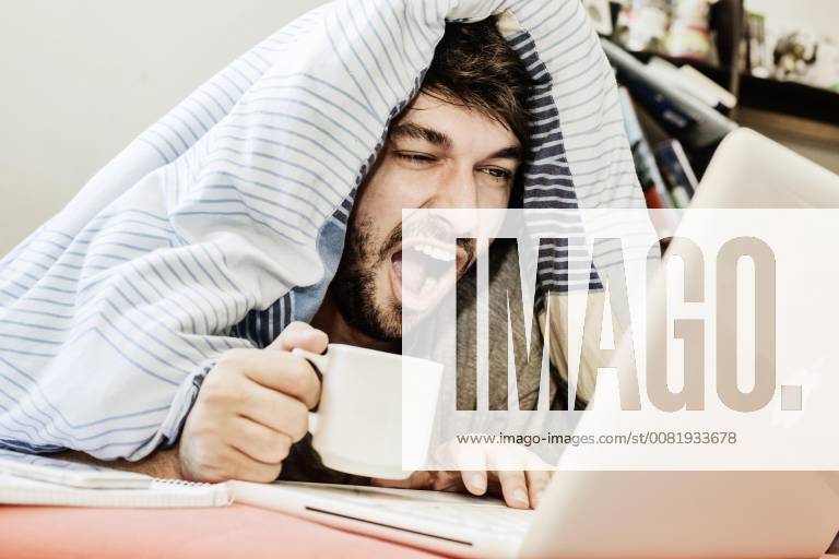 Young man learns Home Office Germany Europe yawning works on a computer in bed
