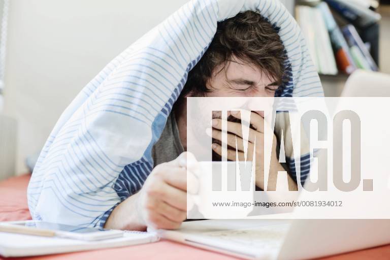 Young man learns Home Office Germany Europe yawning works on a computer in bed