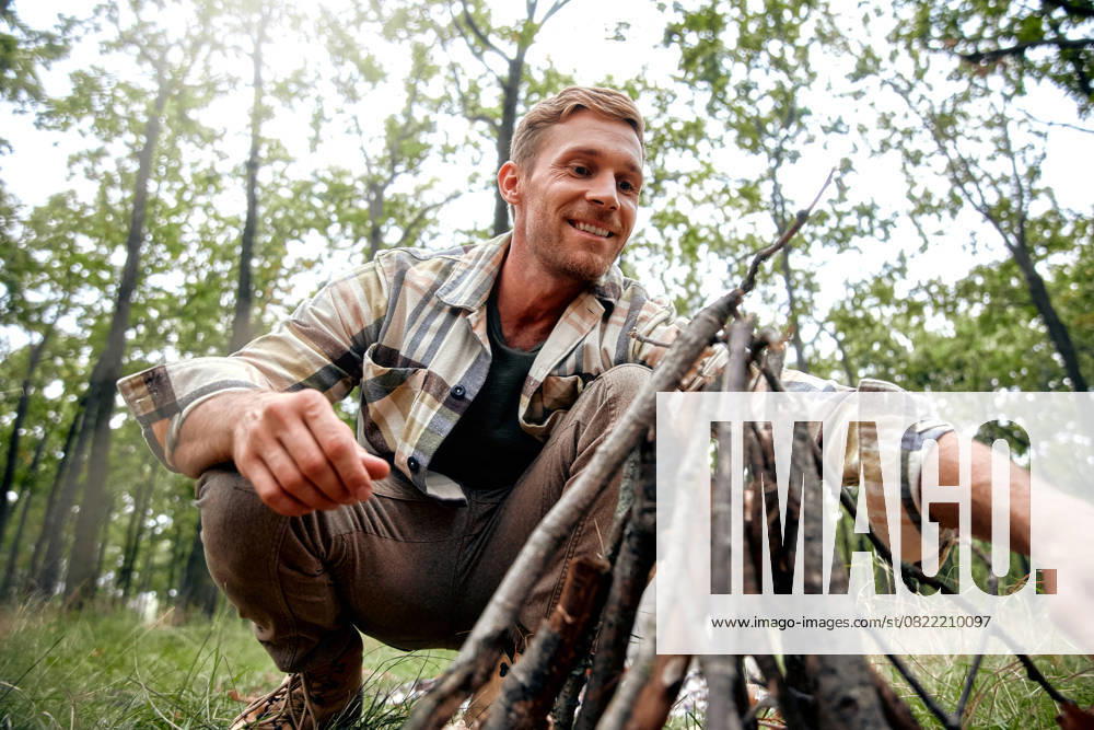 Joyful Outdoorsman Getting Ready to Prepare Campfire in a Beautiful Lush Forest Setting A cheerful man with a warm smile crouches by a flickering campfire, enjoying its warmth and the beauty of the forest, embracing outdoor adventures and reflecting on lifes simple pleasures Model Released OrionProduction_ID27801_798220_052