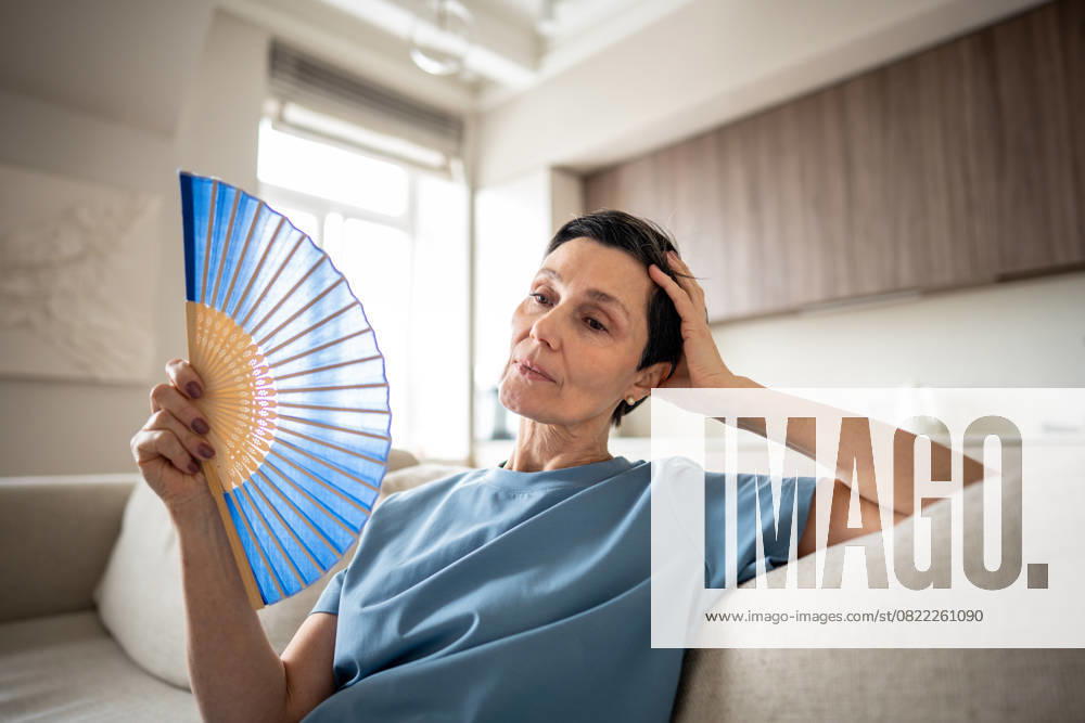 IMAGO Creative, Gesellschaft, Leisure Sickness Erschöpfte ältere Frau, die einen Ventilator benutzt, um sich in einer brütend heißen Wohnung abzukühlen. Hitzeerschöpfung leidet Exhausted mature woman fanning herself at home due to unbearable heat. Elderly suffering from stuffy apartment conditions, discomfort without AC, tropical climate struggle, aging fatigue, menopause (License=RF) ,model released, Symbolfoto